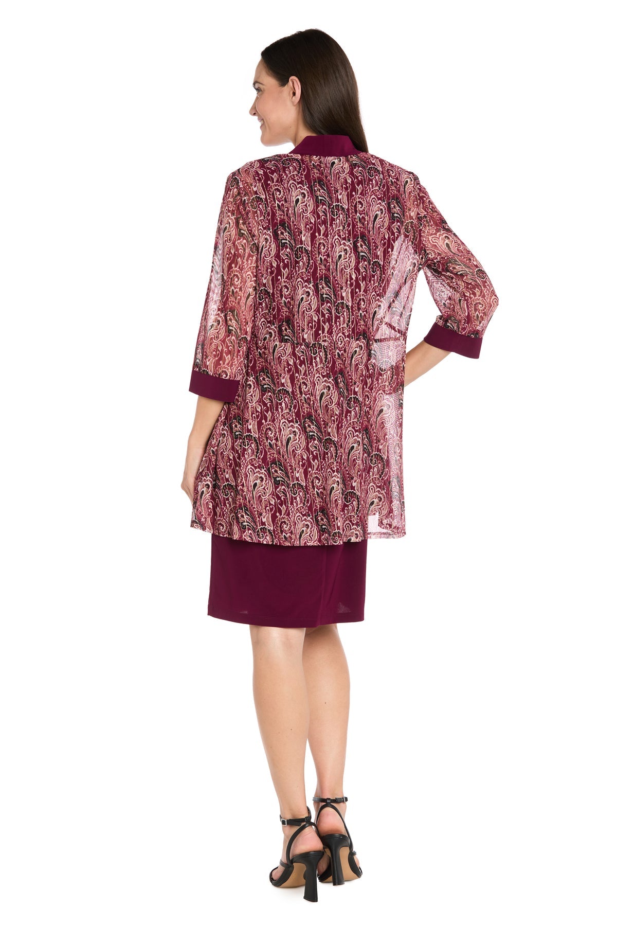A woman stands with her back to the camera in the R&M Richards 3029P Petite Short Wine Party Dress, featuring a knee-length burgundy dress with a matching sheer, patterned jacket and black heeled sandals—ideal for any semi-formal event.